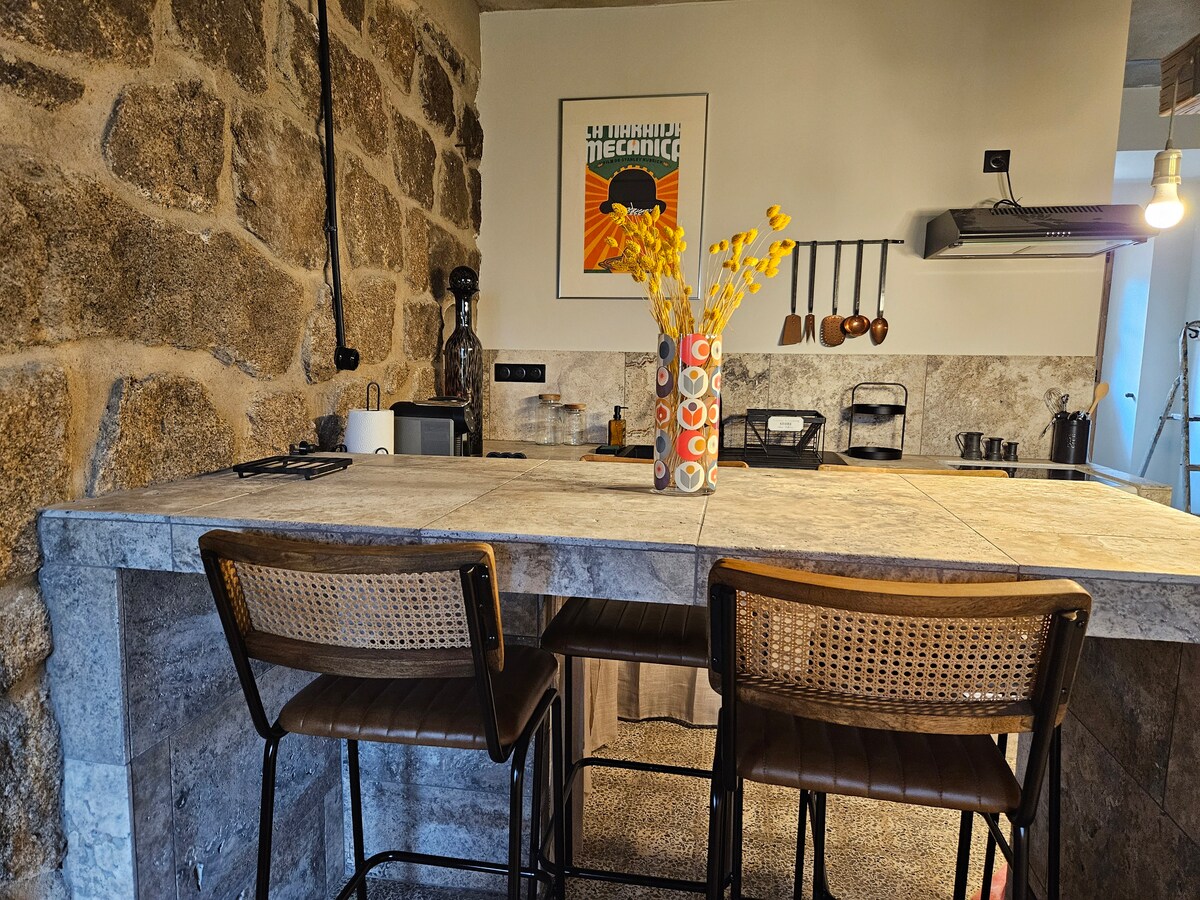 The kitchen area features a stone wall and a modern bar with two tall chairs. A vase with dried flowers adds a touch of decor, while essential appliances, including a coffee maker and kitchen utensils, are visible. The overall setting reflects a blend of rustic and contemporary design.