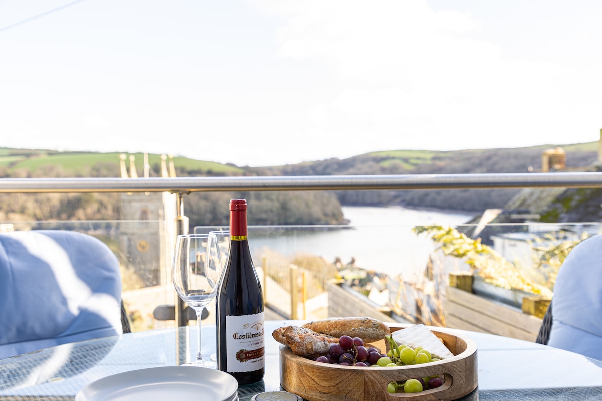 A dining table set for relaxation showcases a bottle of red wine alongside two glasses. A wooden platter presents a selection of grapes, olives, and bread. In the background, panoramic views of the river and lush greenery are visible, enhancing the outdoor dining experience.