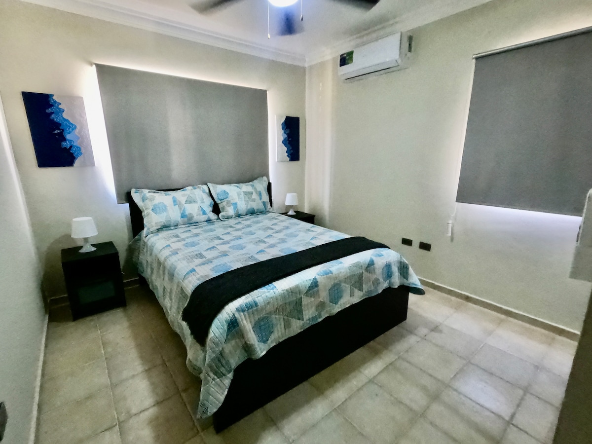 A well-lit bedroom features a spacious bed with a patterned blue and white quilt. Two bedside tables with lamps are positioned on either side. Soft gray shades cover the windows, while a ceiling fan provides additional comfort. Air conditioning unit is mounted on the wall.