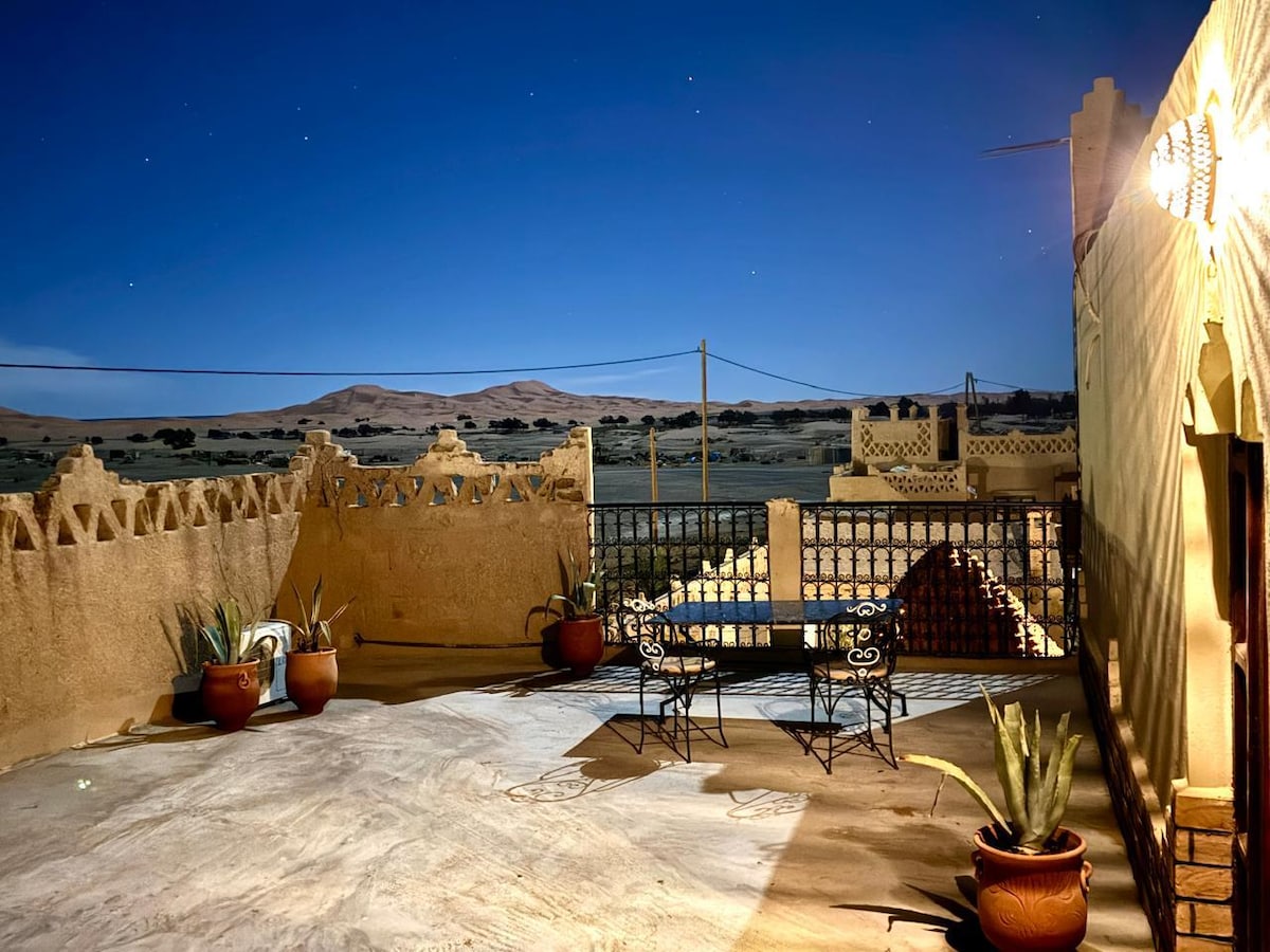 An outdoor terrace features a flat surface with two decorative metal chairs positioned nearby. Potted plants add greenery against the rustic walls. In the background, gently rolling sand dunes are visible under a clear night sky, showcasing the surrounding desert landscape.