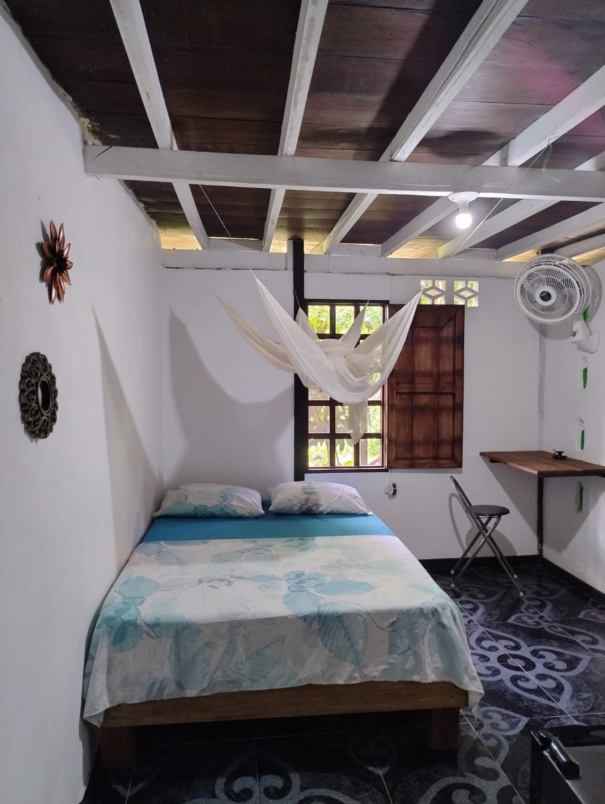 A bright room features a spacious bed draped with a light-colored blanket. Natural light filters through a window adorned with wooden accents. A small folding table and chair are positioned near the wall, while a fan provides air circulation overhead.