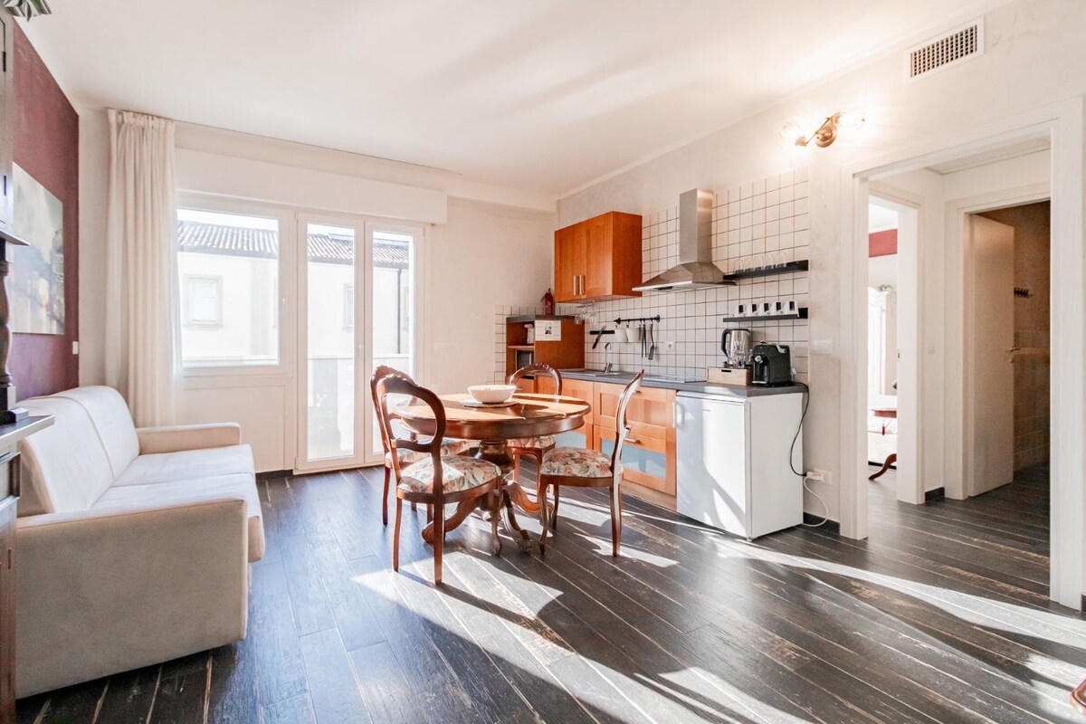 A well-lit living area features a wooden dining table surrounded by four chairs. A cozy sofa is positioned nearby. The kitchen displays modern appliances, including a stovetop, oven, and refrigerator, while natural light filters through the nearby windows.
