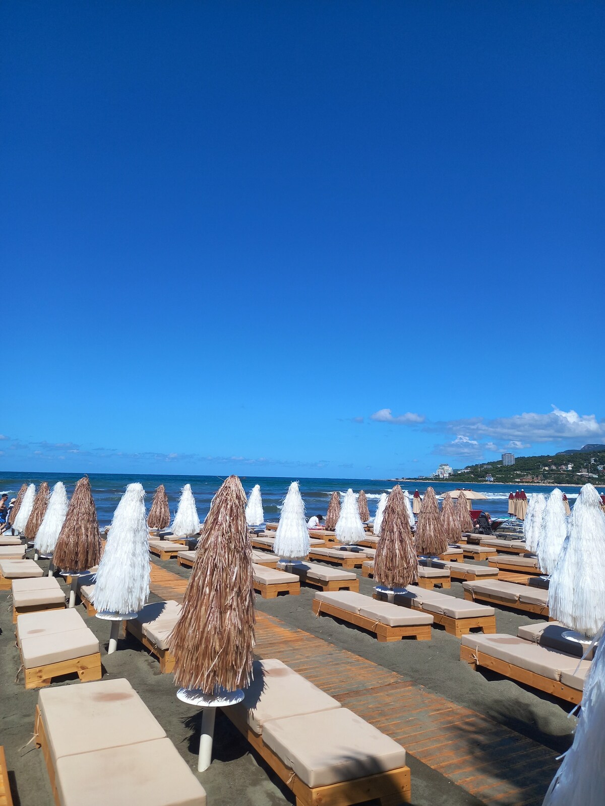 A sandy beach area is filled with wooden lounge beds, each topped with light-colored cushions. Tall, woven umbrellas provide shade, creating a relaxed atmosphere. The clear blue sky stretches overhead, while the gentle waves of the ocean can be seen in the background.