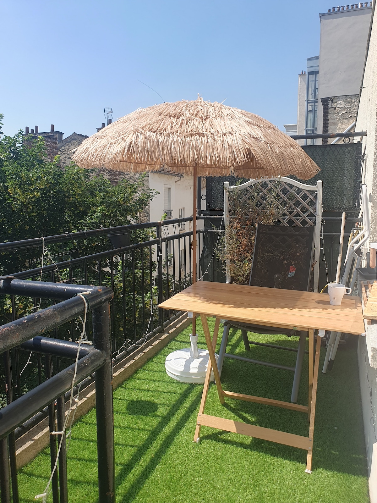 A balcony terrace features artificial grass, a wooden table set for outdoor dining, and a large umbrella providing shade. Lush greenery is visible beyond the railing, enhancing the serene atmosphere. The space offers a cozy area to relax or enjoy meals outside.