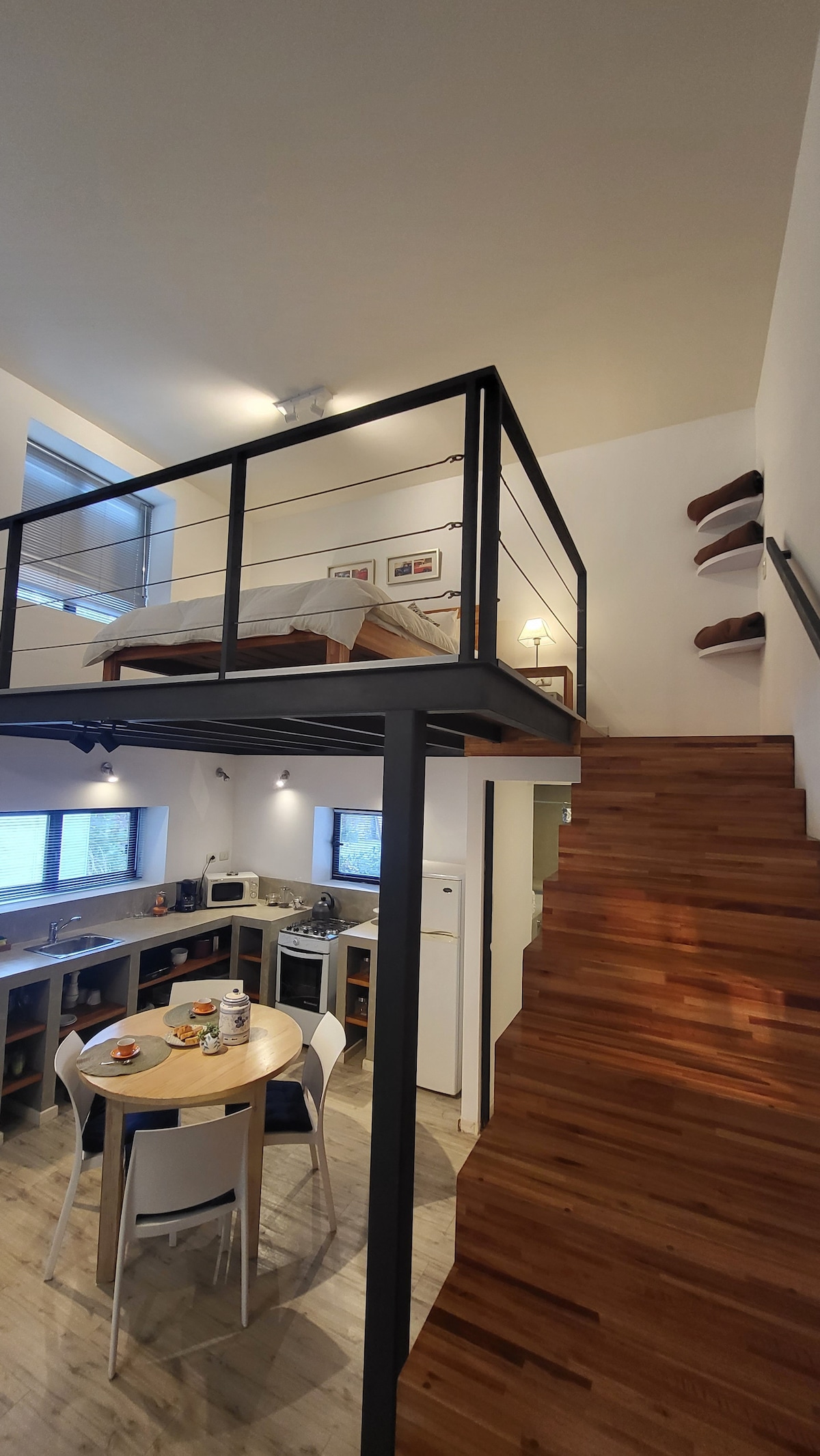 A loft-style interior is shown, featuring a raised sleeping area with a spacious bed visible above. The lower level includes a dining table with chairs, a kitchenette area, and wooden stairs leading to the upper floor. Natural light filters through large windows.