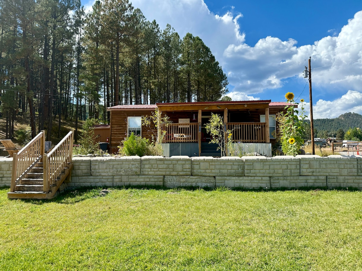 A cozy home is set against a backdrop of tall pine trees, featuring a welcoming front porch with railings. A well-maintained lawn surrounds the property, complemented by a stone wall. Sunflowers add a touch of color near the entrance.