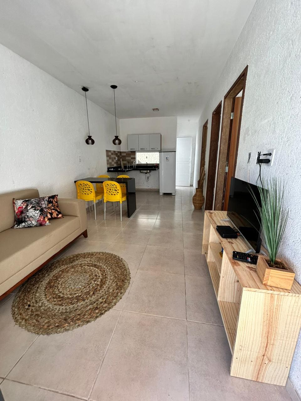 An open living space features a neutral-toned sofa adorned with a decorative pillow, complemented by a round woven rug. A small dining area with yellow stools sits beside a modern kitchen. A television and decorative plant are placed on a wooden console against the wall.