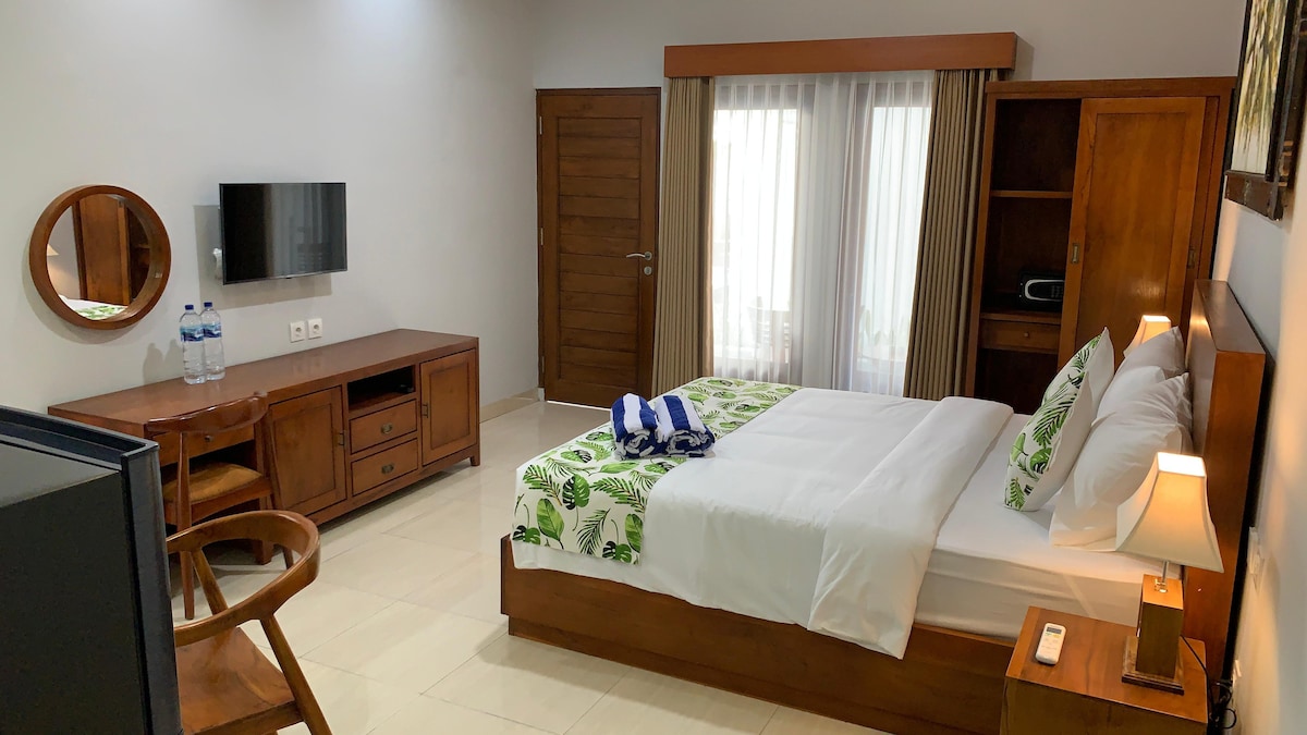 A cozy room features a spacious bed with fresh linen and decorative pillows. A wooden desk and chair are positioned nearby. A television is mounted on the wall, and natural light filters through sheer curtains, illuminating the warm wooden accents in the room.