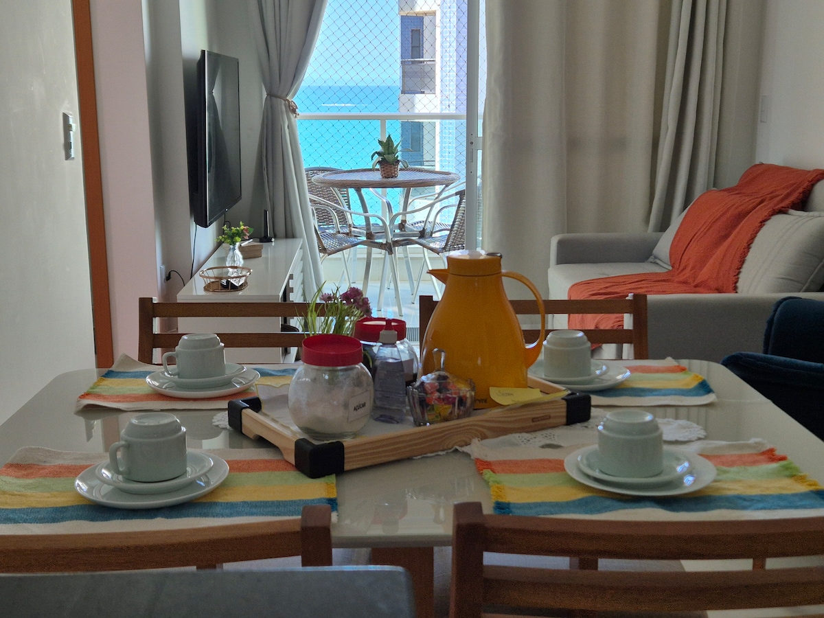 A dining table is set with four light blue cups, accompanied by a jar of sweets and colorful placemats. In the background, a living area is visible, featuring a gray sofa and a balcony with a view of the ocean. Natural light illuminates the space.