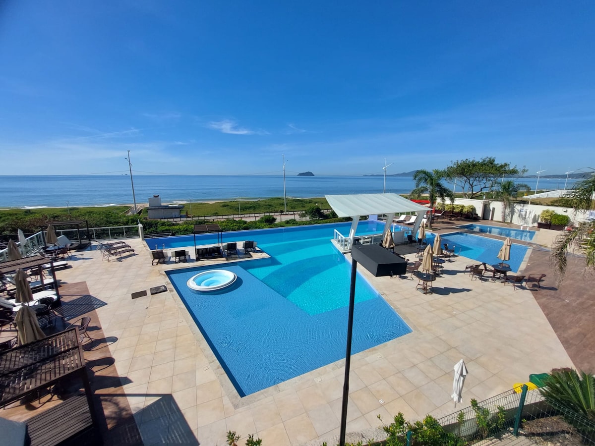 A spacious outdoor pool area is visible, featuring a large swimming pool and a separate hot tub. Lounge chairs are arranged around the pools, with the ocean horizon in the background. Palm trees provide a touch of greenery in the well-maintained landscape.
