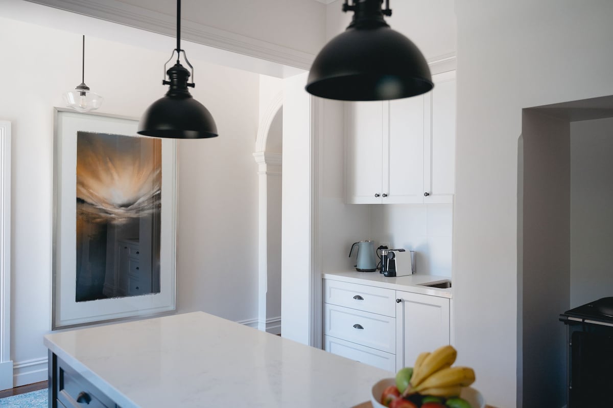 A modern kitchen is displayed, featuring white cabinetry and a central island with a subtle marble surface. Pendant lights provide illumination, and a bowl of fruits sits on the island. An artwork in muted tones adds a decorative touch to the space.