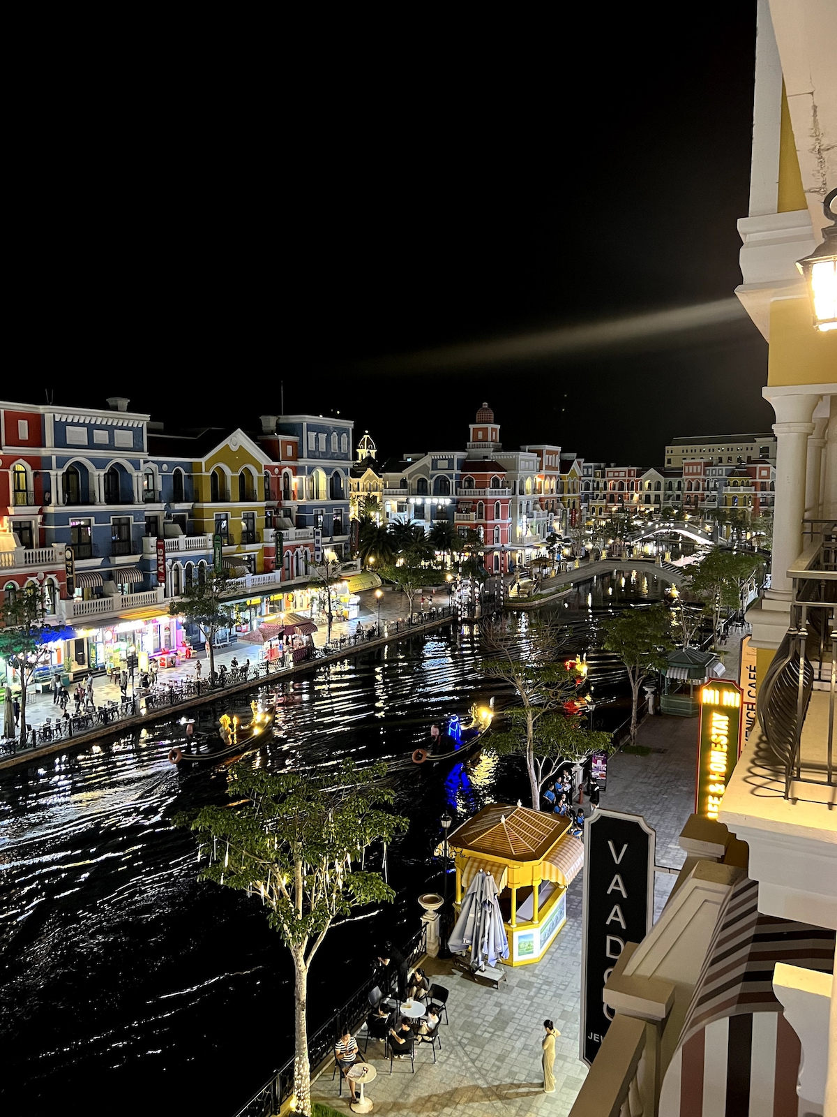 An aerial view captures a vibrant night scene along a canal, illuminated by colorful buildings reflecting in the water. Strolling pedestrians and seated diners are visible, with trees lining the walkway and a gazebo adding character to the lively atmosphere.
