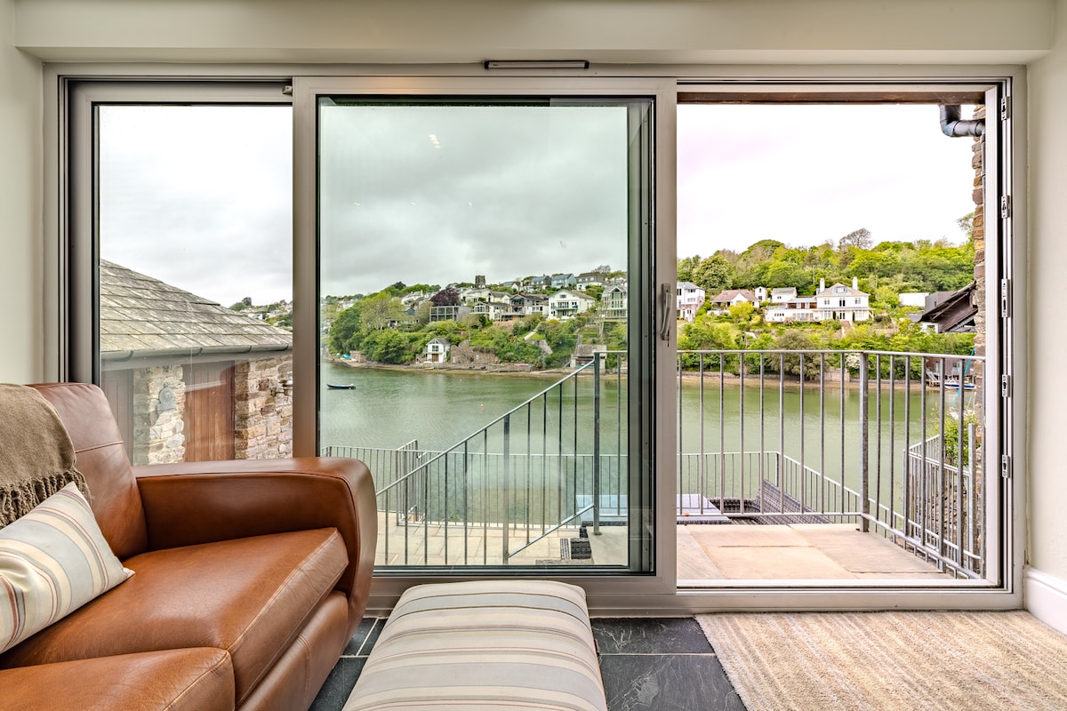 A large floor-to-ceiling glass wall showcases views of the River Yealm, with a calm water surface reflecting the surrounding greenery. A cozy seating area with leather sofas is positioned nearby, providing a comfortable spot to enjoy the scenery through the sliding doors.