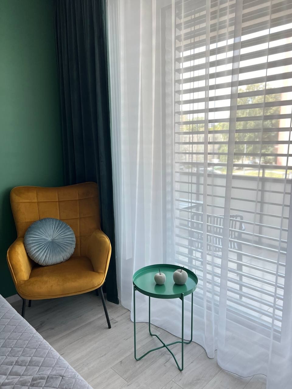 A cozy seating area features a bright yellow armchair with a decorative pillow. A green side table holds two small decorative items. Sheer curtains diffuse natural light coming through the window, offering a view of the outside while maintaining privacy.