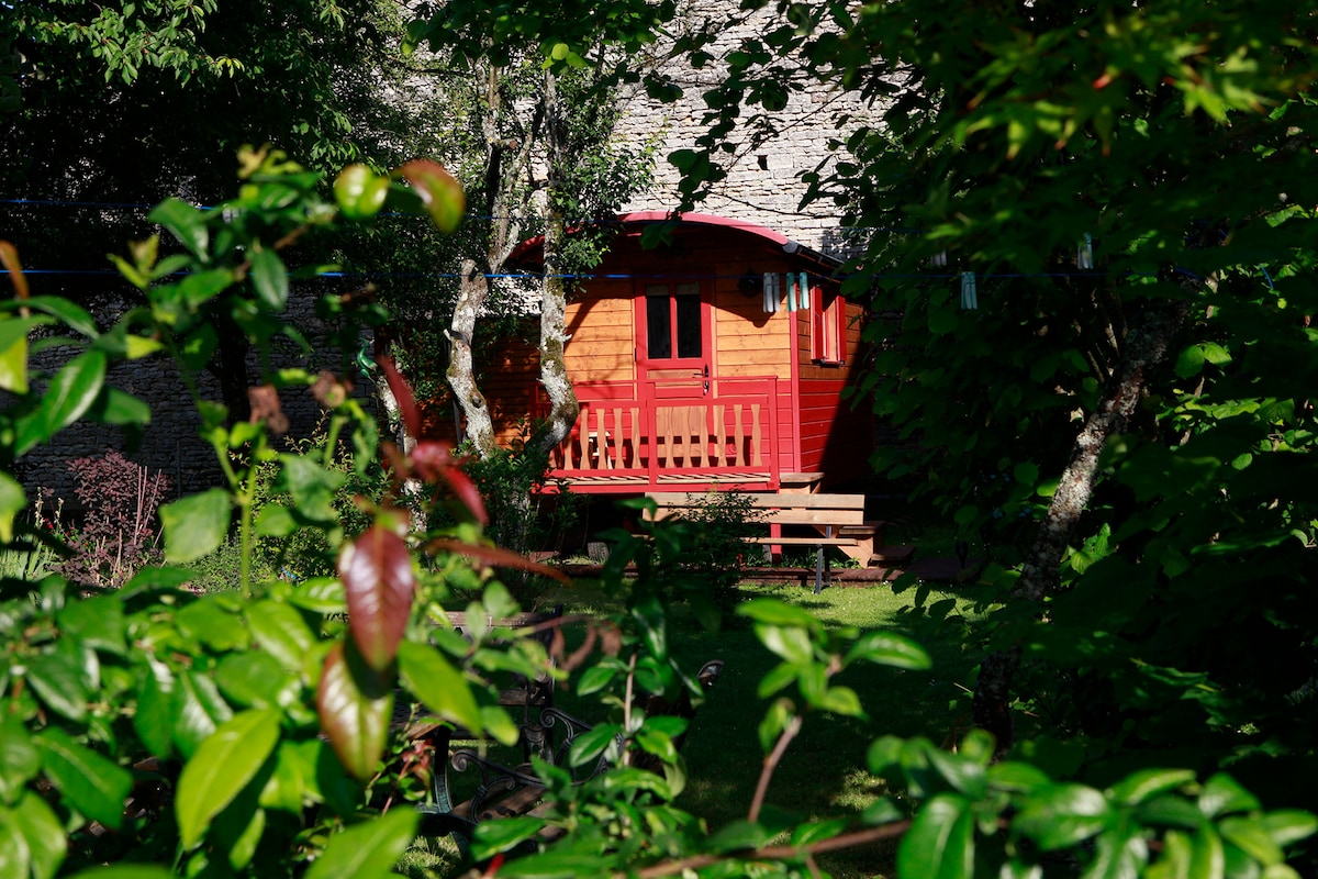 The rustic exterior of a red and wooden roulotte is visible through lush greenery. A small porch with seating overlooks a well-maintained garden, providing an inviting and serene outdoor space amidst the surrounding nature.