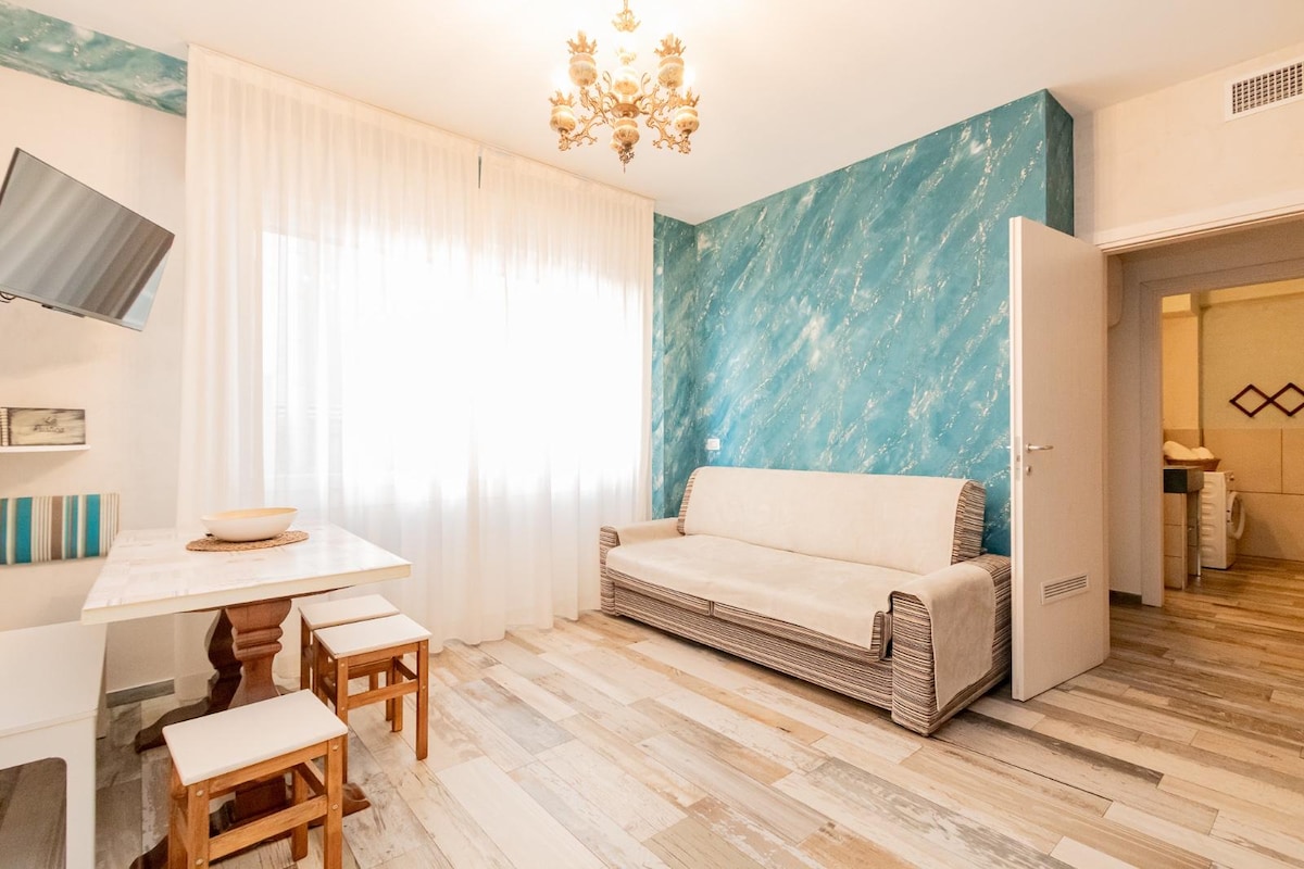 A light-filled living area features a cozy sofa, a dining table with chairs, and a mounted television. Textured blue walls and a chandelier add visual interest. The space is complemented by a doorway leading to a well-appointed bathroom.