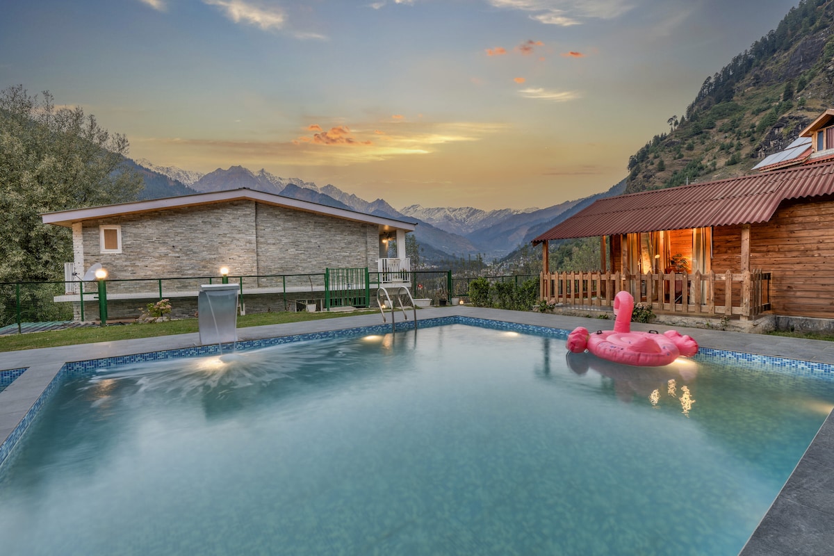 A serene outdoor pool area is surrounded by lush greenery and mountains. Comfortable seating is visible near the pool, and an inflatable flamingo floats on the water. The sunset casts a warm glow over the scene, enhancing the natural beauty of the setting.