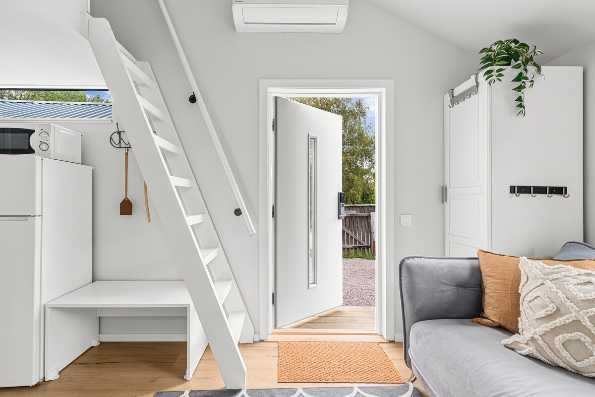 A bright entrance area features a white door leading outside, complemented by a ladder ascending to the loft. A modern couch is positioned nearby, with a decorative cushion on display. A compact kitchen area is visible, along with a small dining space.