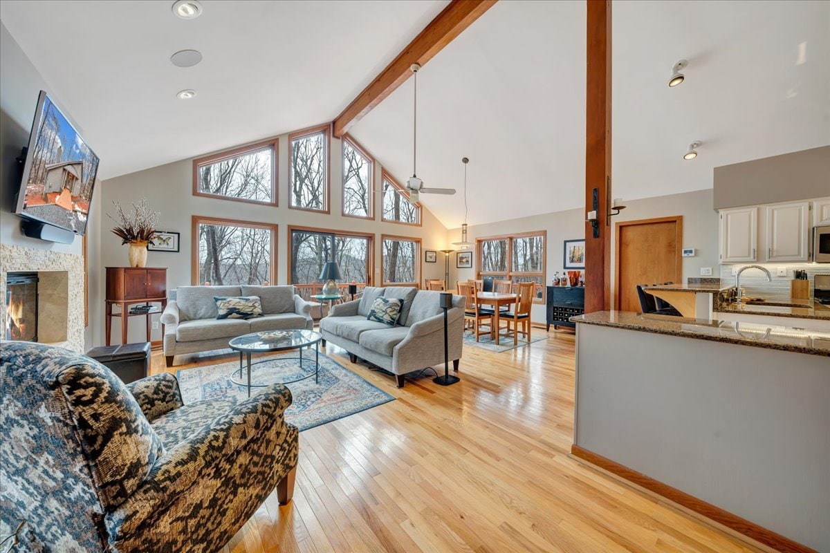 An open living space features large floor-to-ceiling windows that provide ample natural light. Two comfortable sofas and a patterned armchair surround a glass coffee table, while a dining area with a wooden table is visible in the background. A flat-screen TV is mounted above a modern fireplace.