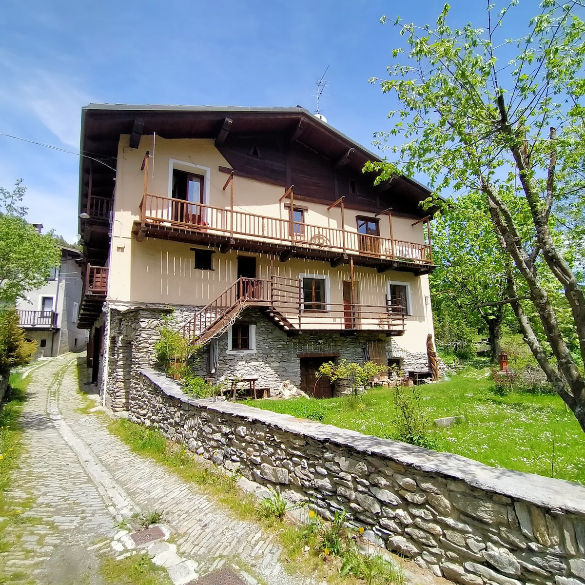 A three-story house is set amidst greenery, featuring a stone wall and a pathway leading to the entrance. Balconies are visible on the upper floors, and windows allow natural light to illuminate the exterior. Trees and grass surround the structure, enhancing the natural environment.