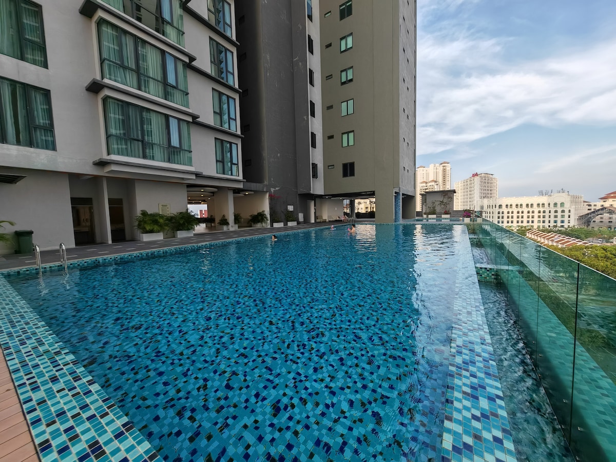 A large swimming pool is visible, featuring a vibrant blue mosaic tile design. Clear glass panels border the pool area, offering unobstructed views of the surrounding skyline and greenery. The atmosphere is peaceful, complemented by well-maintained landscaping along the pool's edge.