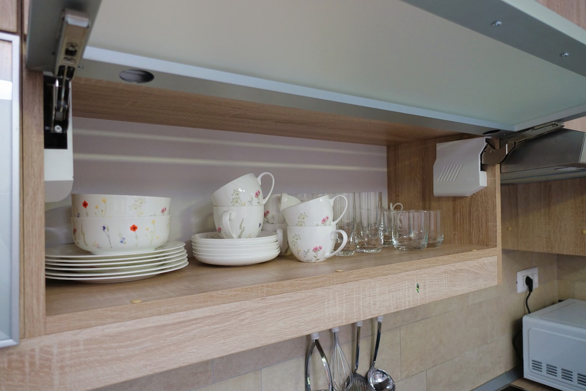 A kitchen shelf displays neatly arranged dishware, including several bowls and plates alongside stacks of cups. Glasses are positioned at the back, showcasing a variety of shapes. The overall organization emphasizes functionality and convenience for meal preparation.