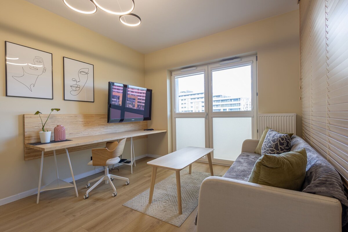 A modern living area features a comfortable sofa adorned with cushions. A wooden desk with a chair is positioned adjacent to the large television, while natural light enters through two glass doors leading to a small balcony. Minimalist artwork is displayed on the walls.