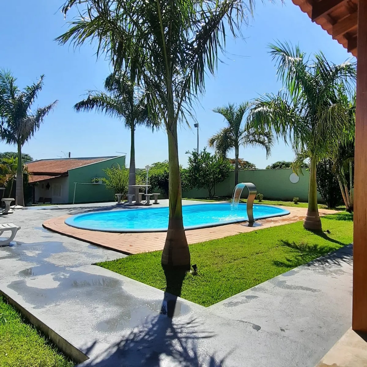 The outdoor area features a spacious swimming pool surrounded by palm trees and well-maintained grass. Adjacent seating areas are visible, providing a relaxing space for guests. Clear blue skies and sunlight enhance the inviting atmosphere of the landscaped yard.