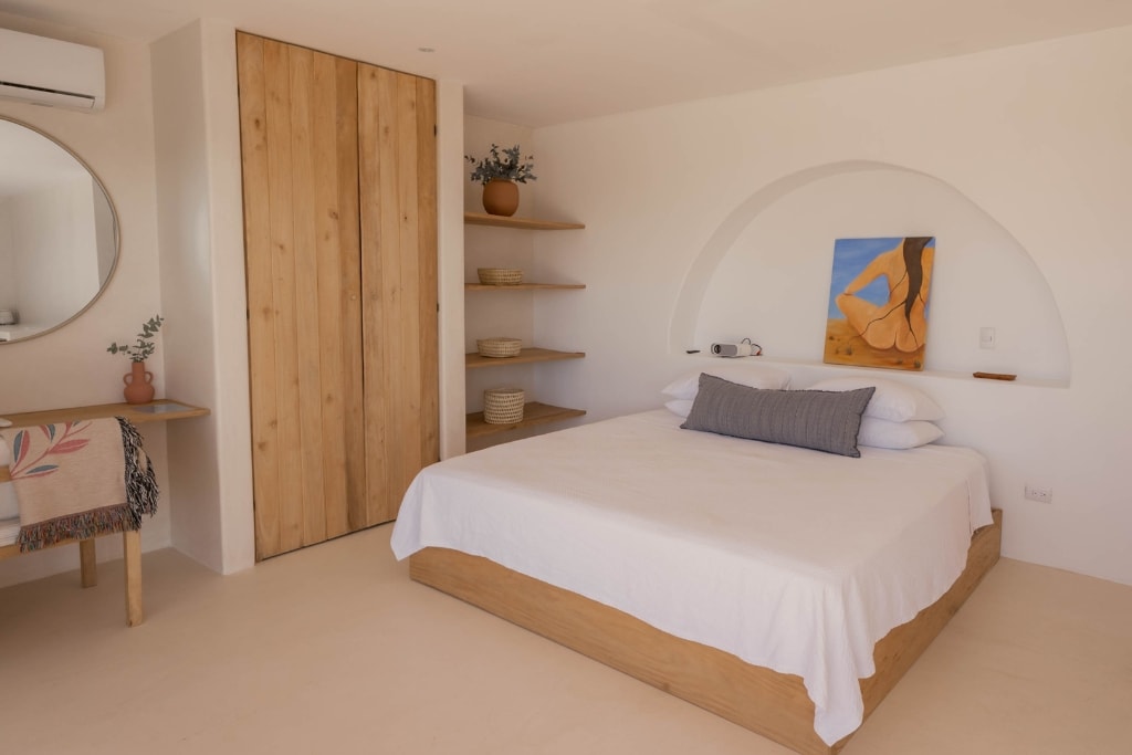 A calming sleeping area features a spacious bed with a light-colored bedspread. Soft natural wood accents are found in the bed frame and a nearby shelf. A round mirror and a small table are included, adding functional elegance to the room.