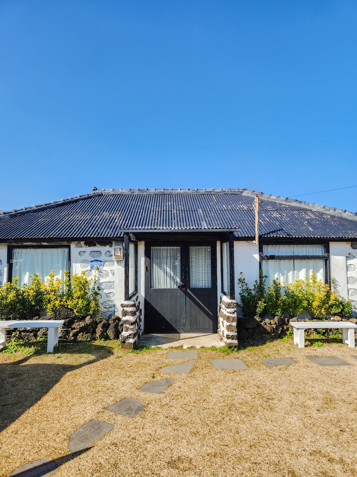 A charming traditional stone house features a thatched roof and a center black door flanked by large windows. Vibrant yellow flowers adorn the exterior, while stone pathways lead to the entrance. The clear blue sky provides a bright backdrop to the inviting façade.