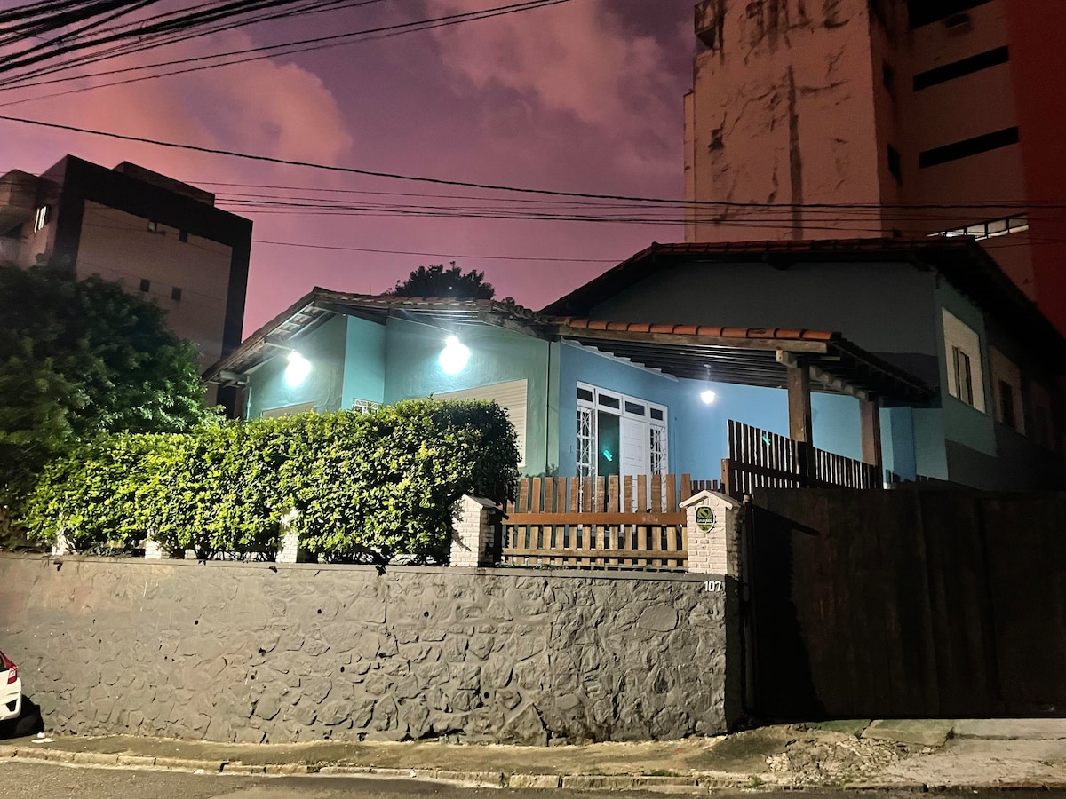 The exterior of a charming house is illuminated at dusk, showcasing a light blue facade and a sloped roof. Lush greenery surrounds the property, complemented by a low stone wall. Soft lighting highlights the entrance, creating an inviting atmosphere amidst the urban setting.