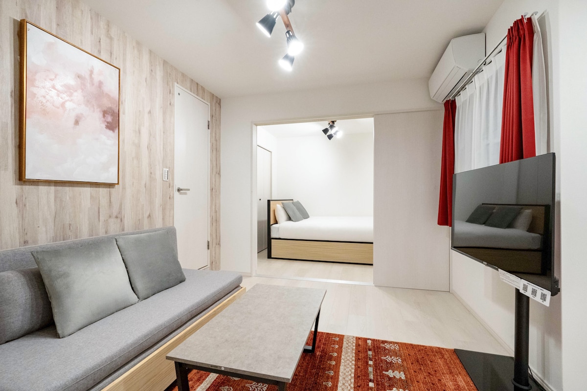 An open living area features a gray sofa with two accent pillows and a low coffee table, complemented by a colorful rug. A wall art piece in soft tones is displayed above. A partition leads to a modest bedroom area with a spacious bed, softly illuminated by ceiling lights.