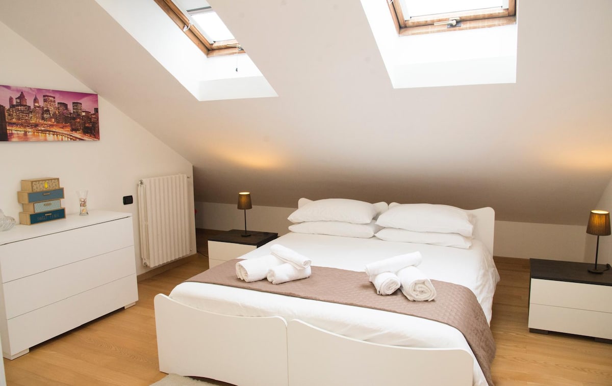 A bright bedroom features a spacious bed with white linens and a brown throw. Two bedside lamps provide soft illumination, and a chest of drawers offers additional storage. Skylights bring in natural light, enhancing the room's airy ambiance.