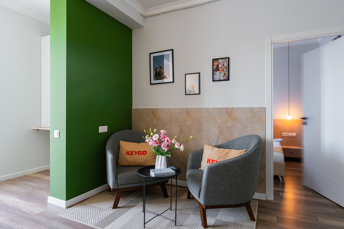 A small seating area features two gray armchairs adorned with branded pillows. A round black table sits between them, complemented by a floral arrangement. The wall exhibits framed pictures, and a stylish green wall adds a modern touch to the space.