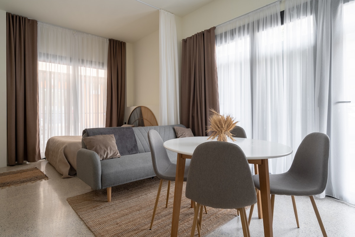A modern living area is presented with a gray sofa and a circular dining table surrounded by four gray chairs. Natural light filters through sheer curtains, illuminating the space. A simple decorative arrangement is placed in the center of the table, enhancing the clean and relaxed atmosphere.