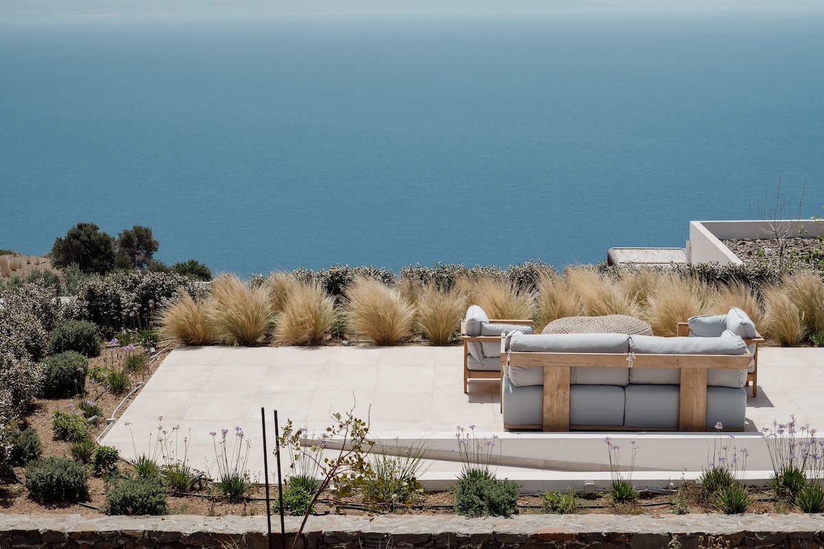An outdoor seating area features a spacious lounge sofa, positioned to offer unobstructed views of the crystal blue sea. Surrounding the area, soft coastal grasses and flowering plants add natural beauty to the terrace, which is designed for relaxation and enjoyment of the scenery.