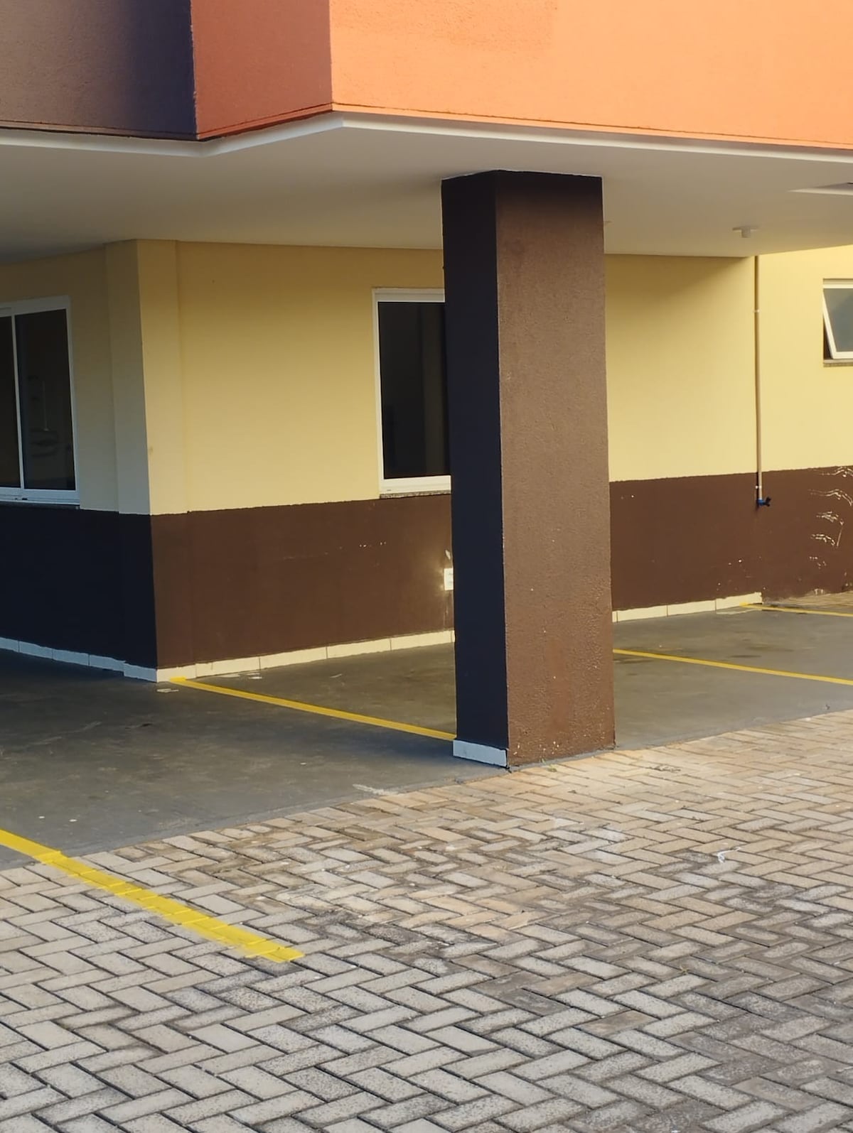 A portion of the building exterior is shown, featuring a solid structural column at the corner. The textured pavement is visible, showcasing marked parking spaces bordered by a brown and cream two-toned wall. Two windows provide natural light to the enclosed area.