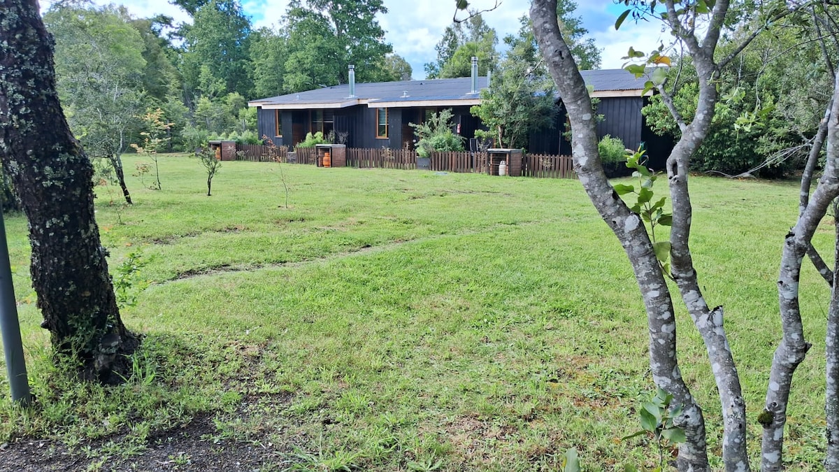 A single-story wooden cabin is nestled in a green landscape, surrounded by trees. A fenced area can be seen in the foreground, leading to a well-maintained lawn. The cabin features multiple windows, allowing natural light to illuminate the interior.
