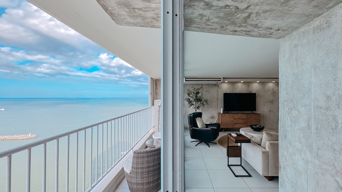 A spacious living area is visible, showcasing a modern design with a large smart TV mounted on the wall. Large glass sliding doors lead to a private balcony, offering unobstructed views of the ocean and sky, enhancing the bright, airy ambiance.