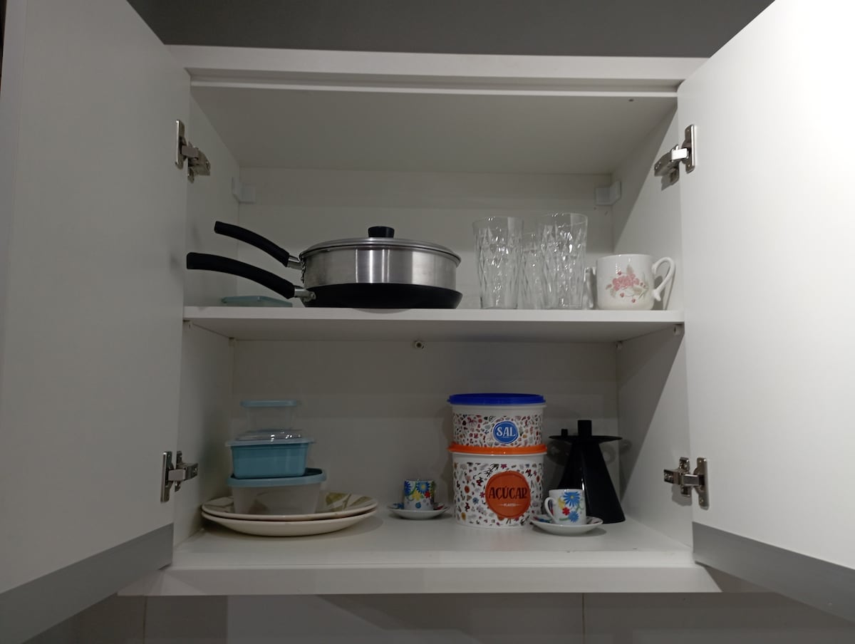 The open kitchen cabinet displays a variety of kitchenware, including a pot, glasses, and a mug. A set of colorful dishes is arranged on the lower shelf, along with containers and a coffee maker, providing essentials for meal preparation.