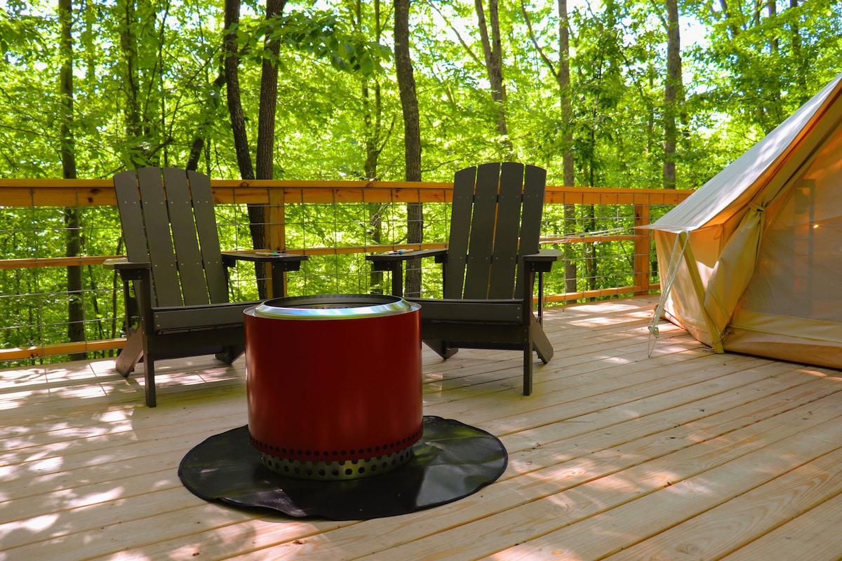 A spacious wooden deck is surrounded by lush green trees, featuring two Adirondack chairs positioned beside a smokeless fire pit. The bell tent is visible in the background, blending harmoniously with the natural setting.