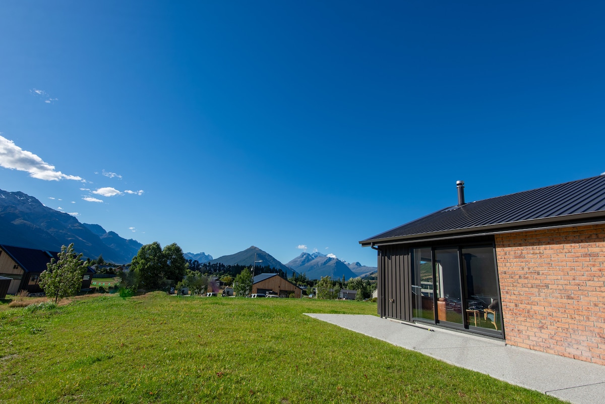 A modern house is set against a backdrop of expansive mountain views. The clear blue sky enhances the landscape, while a landscaped lawn provides an inviting outdoor space. The building features a combination of brick and metal siding, contributing to its contemporary design.