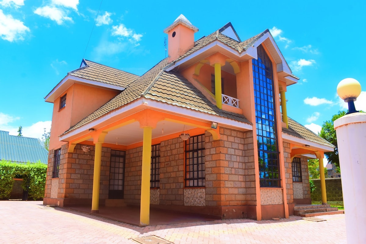 A two-story home is presented with a combination of stone and bright orange walls. Large windows reflect sunlight, and the spacious porch is supported by yellow columns. A tiled walkway leads to the entrance, framed by greenery and clear blue skies.