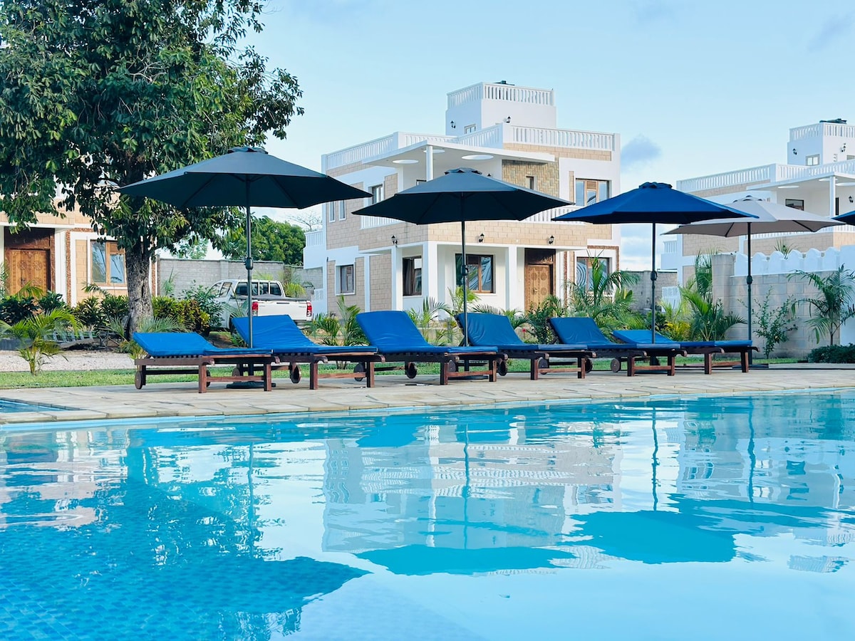 A serene swimming pool reflects the blue sky, surrounded by sun loungers under large umbrellas. The villas are situated in the background, showcasing a light-toned exterior, with green foliage enhancing the tranquil setting.