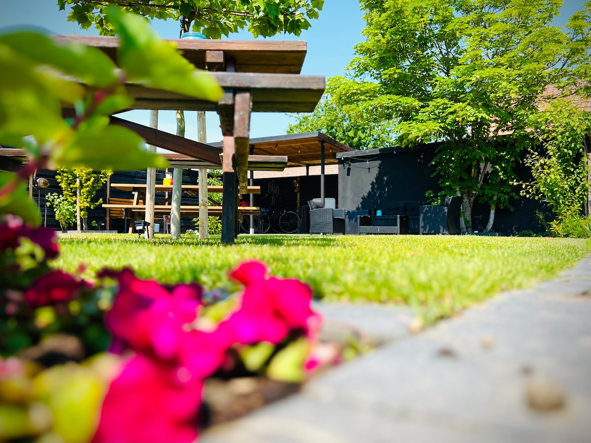 A well-maintained garden is presented, featuring various seating arrangements under a shaded area. The lush green lawn is bordered by vibrant flowers and trees, contributing to a peaceful outdoor environment. A few cycling racks are also visible, enhancing the space's functionality.