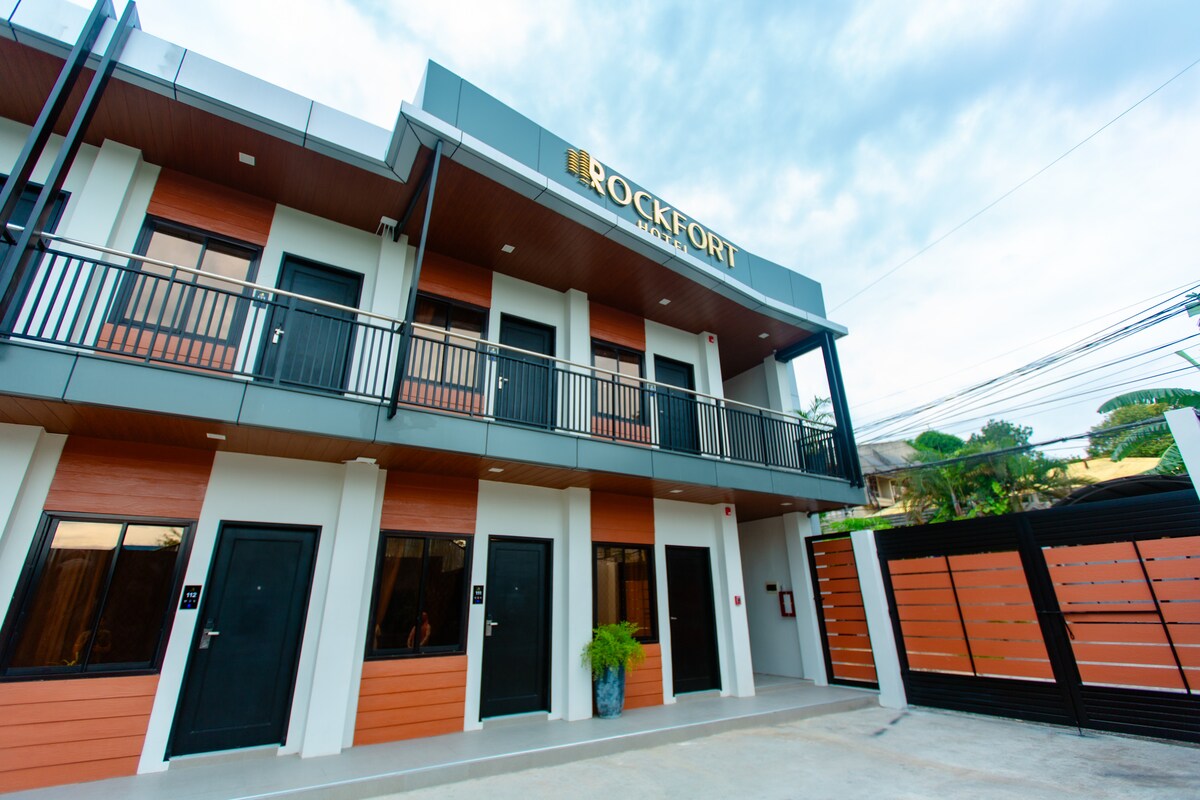 A modern exterior is showcased, featuring two levels of sleek architectural design. The building is highlighted by a prominent sign stating 'ROCKFORT'. Black doors with geometric accents are contrasted against warm wooden panels, complemented by potted plants enhancing the entrance.