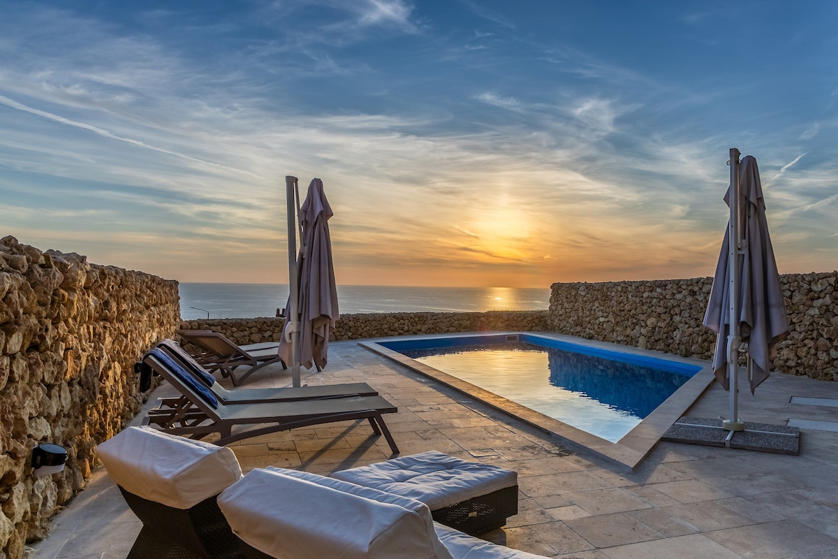 The pool area features a generously sized pool surrounded by stylish lounge chairs and umbrellas. Natural stone walls provide privacy, while the sunset casts a warm glow over the tranquil scene, reflecting off the water.