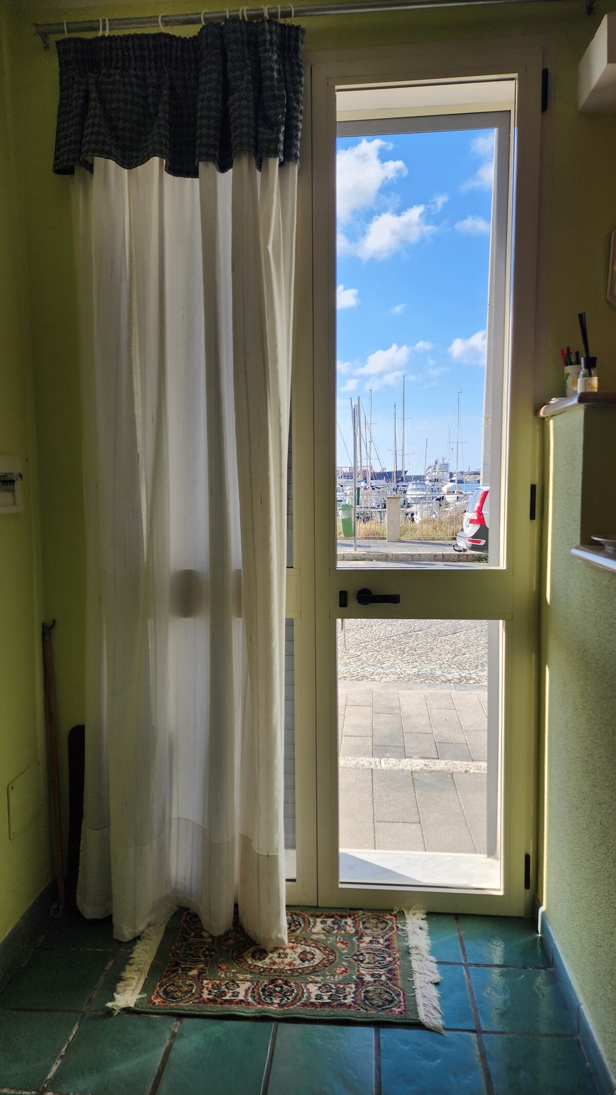 A doorway with a white curtain partially drawn open reveals a scenic view of a marina. The blue sky is dotted with white clouds above the moored boats. The floor is tiled in dark green, complemented by a patterned rug at the entrance.
