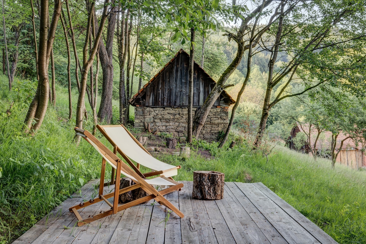 A wooden patio area features a reclining chair made of light-colored wood and a small log as a side table. The background showcases a stone building with a wooden roof, surrounded by lush greenery and trees, offering a serene, natural setting.