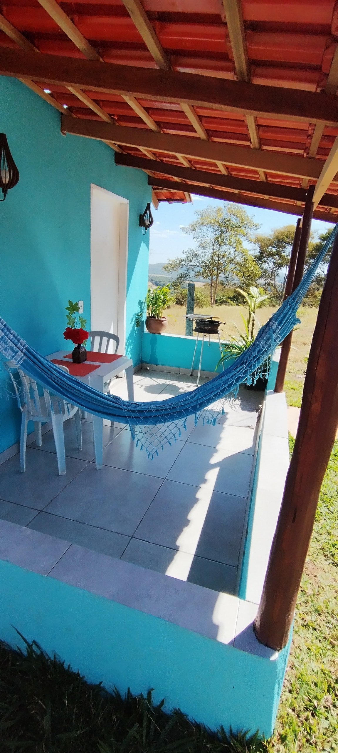 A welcoming porch features a blue hammock draped between posts, accompanied by two white chairs and a small table with a flower vase. The tiled floor extends outdoors, with a lush green landscape visible in the background.